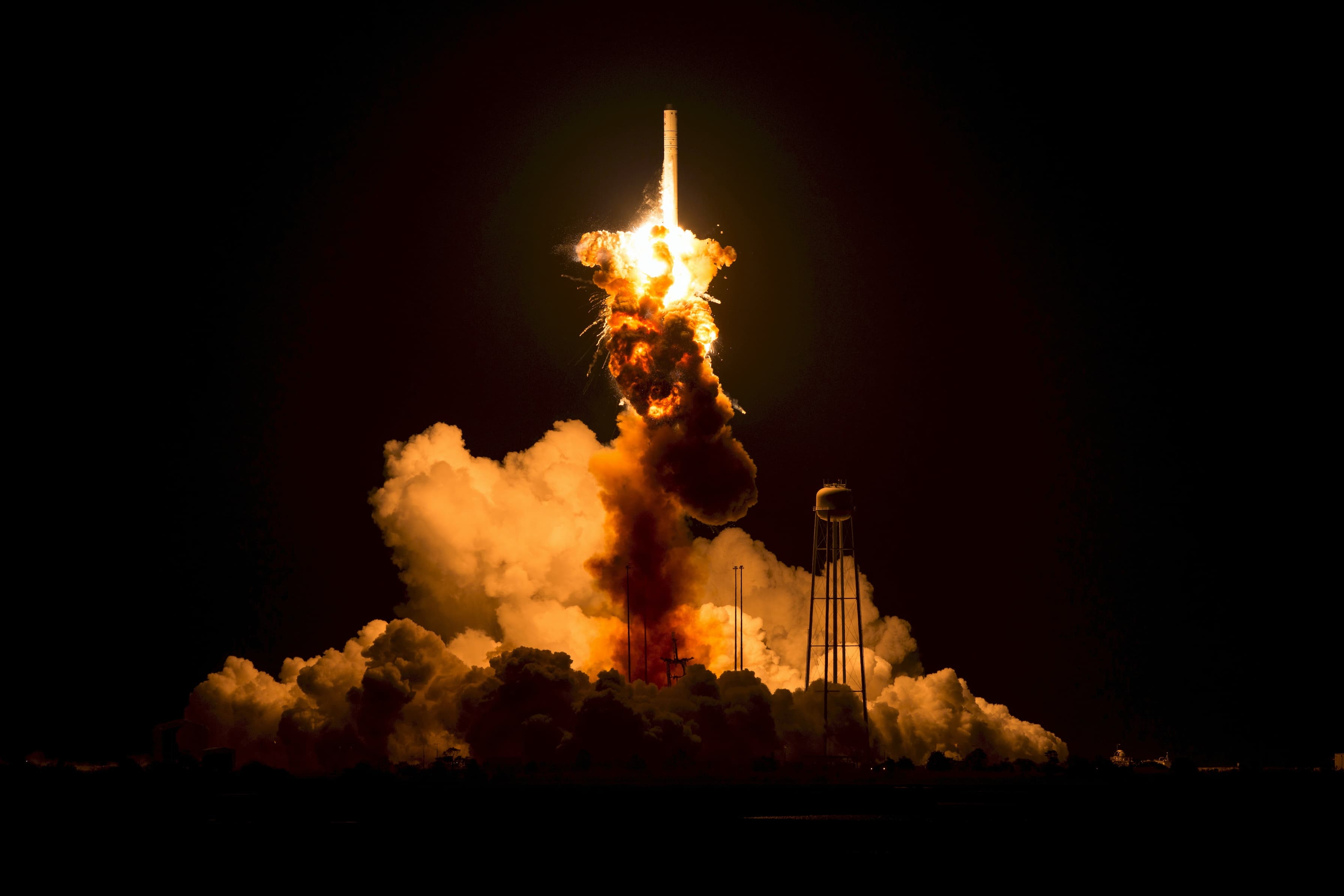 A rocket launching at night with a few small poles and a tower with a tank on top sticking out of a cloud of smoke.